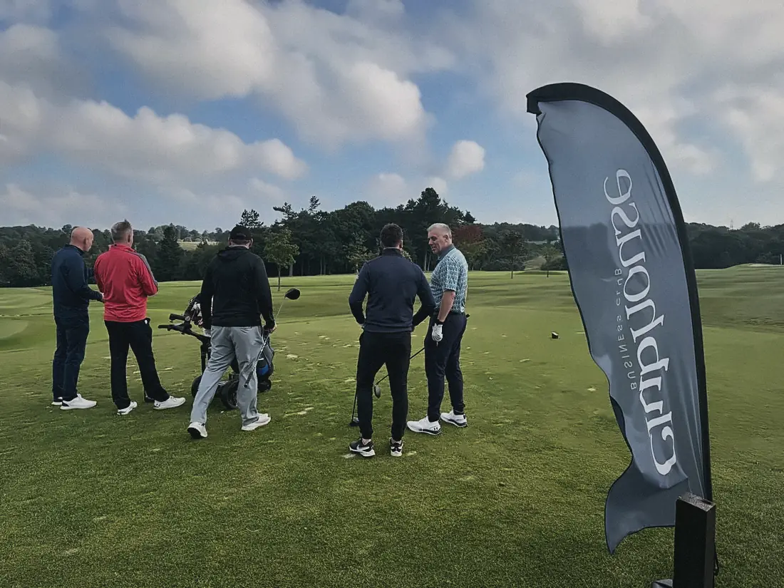 Players gathering by a Clubhouse flag before teeing off