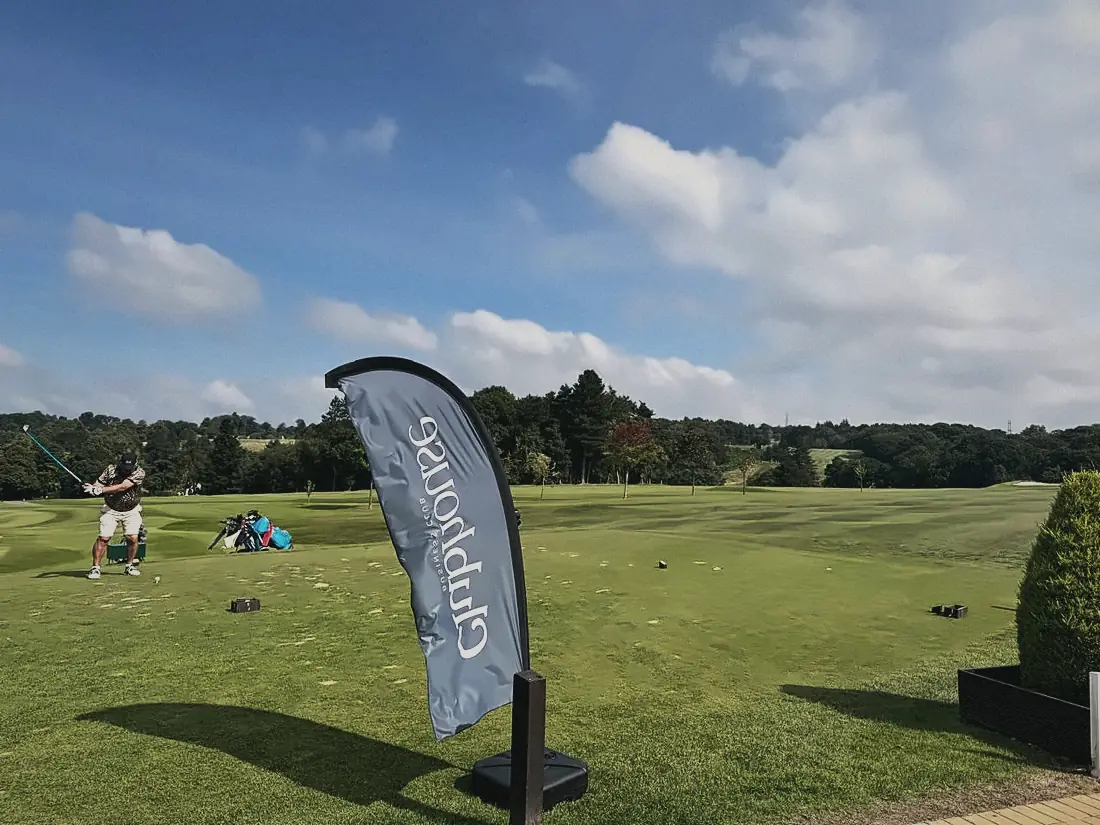 Players on the tee beside a Clubhouse flag under a blue sky